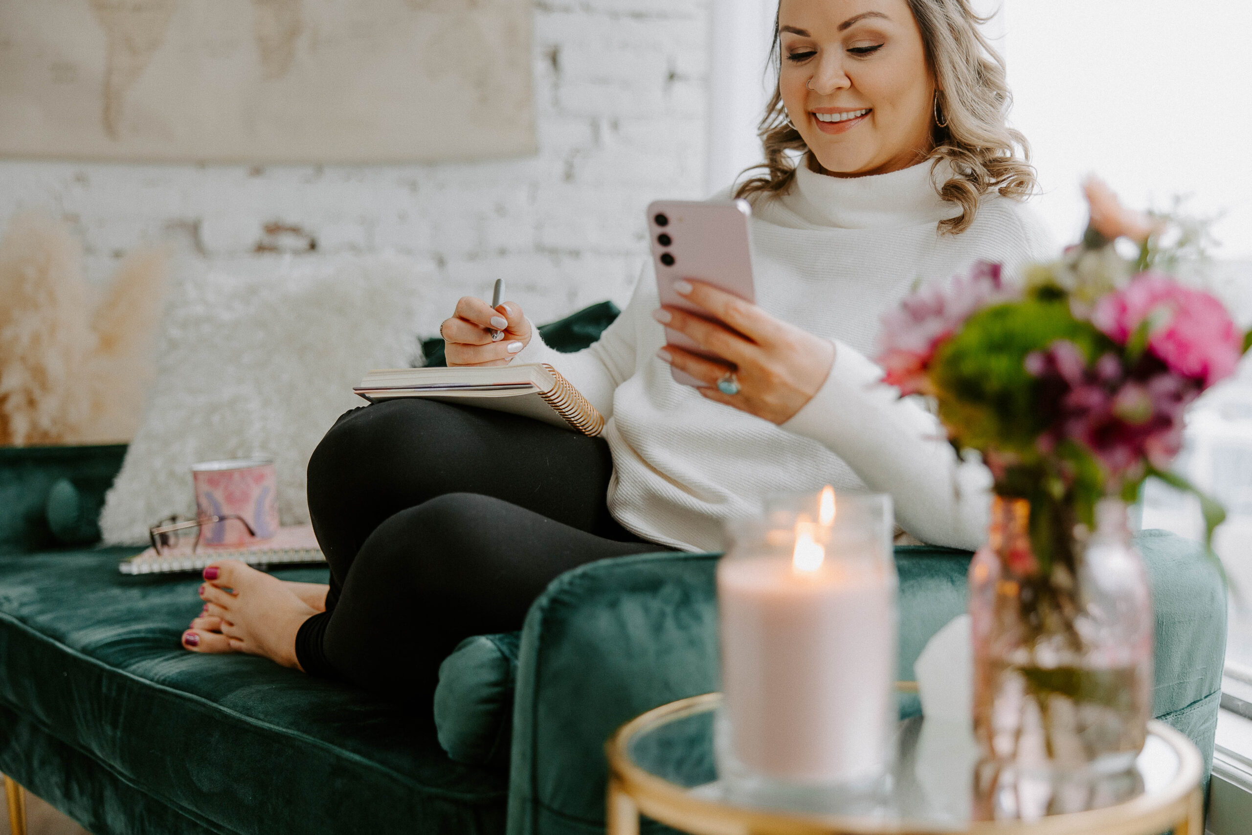 photo of woman sitting on the couch staring at her phone smiling while writing in a notebook