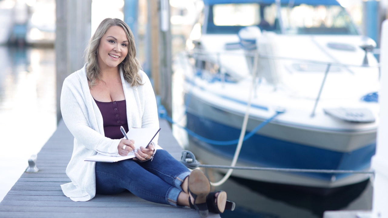 photo of woman sitting on dock with boat behind her and writing in a notebook