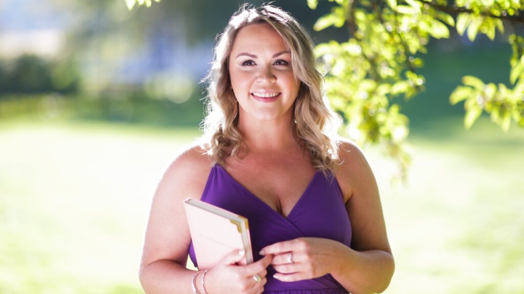 photo of woman standing under tree and smiling while holding a notebook and looking off in the distance